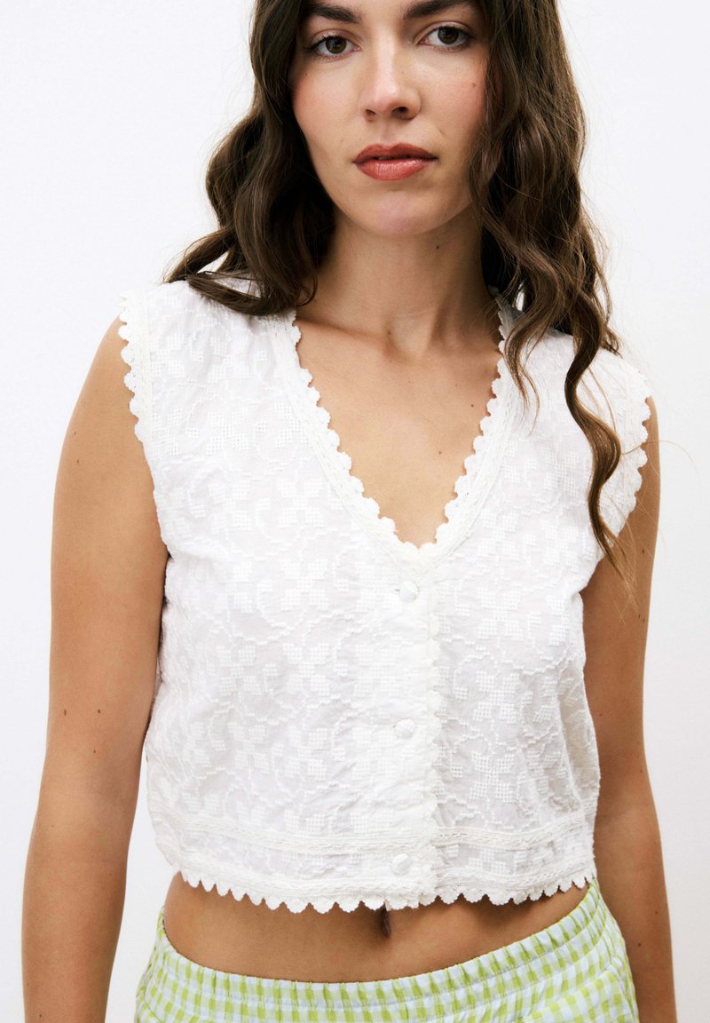 Top court en dentelle blanche avec bords festonnés, décolleté en V et fermeture à boutons à l'avant. Sans manches, présentant un design texturé floral.
