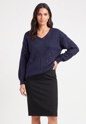 Femme aux longs cheveux foncés portant de grandes boucles d’oreilles anneaux, un pull bleu marine à motif et une jupe noire mi-longue, posant avec une main sur la hanche.