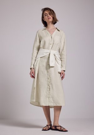 Leinenhemdkleid in hellem Beige mit Knopfleiste, seitlichen Schlitzen und Brusttasche, kombiniert mit dunkelbraunen Riemchensandalen.