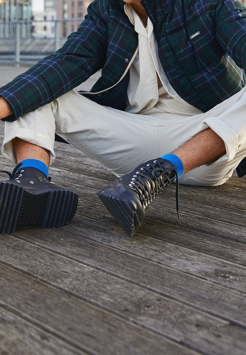 Personne assise sur une terrasse en bois, portant un pantalon beige, des chaussettes bleues, des bottes noires à lacets et une veste à carreaux vert foncé.