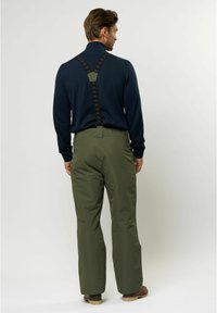 Zielone wodoodporne spodnie z regulowanymi czarnymi szelkami, w połączeniu z ciemnoniebieską koszulą z długim rękawem, widziane od tyłu.