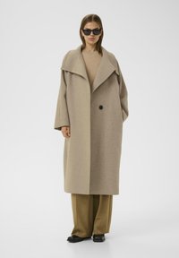 Cappotto di lana beige con collare oversize, chiusura a un bottone nero, maniche lunghe e vestibilità ampia, abbinato a pantaloni marroni e scarpe nere.