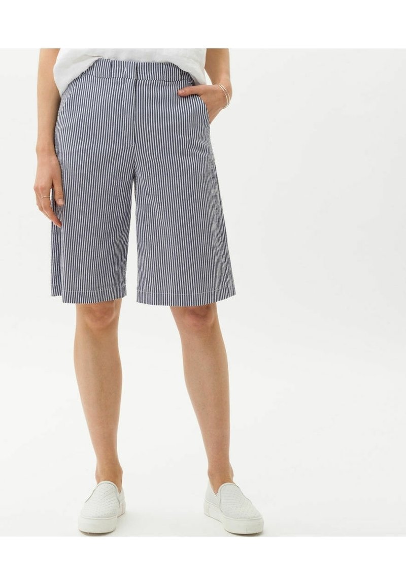 Personne portant un short rayé vertical bleu et blanc arrivant aux genoux avec des poches et des chaussures blanches à enfiler sur un fond blanc.