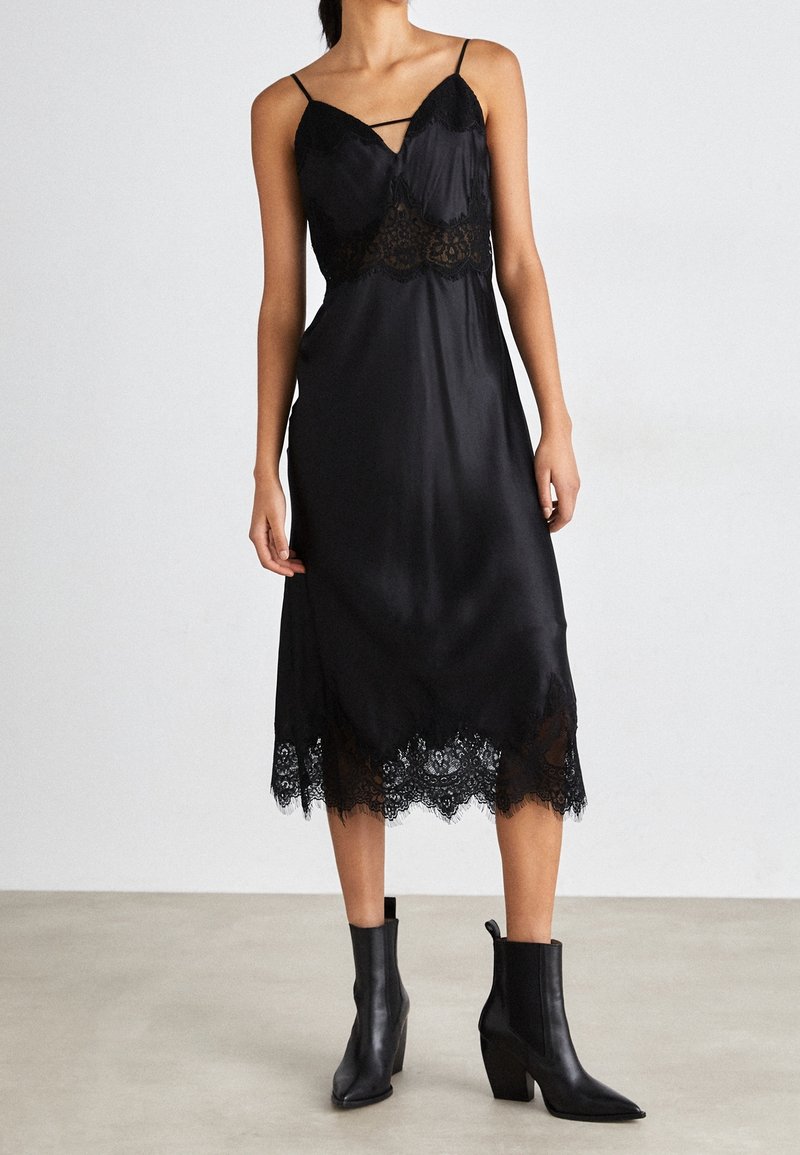 Femme portant une robe nuisette en satin noir avec bordure en dentelle, associée à des bottines noires à bout pointu, debout sur un sol uni.