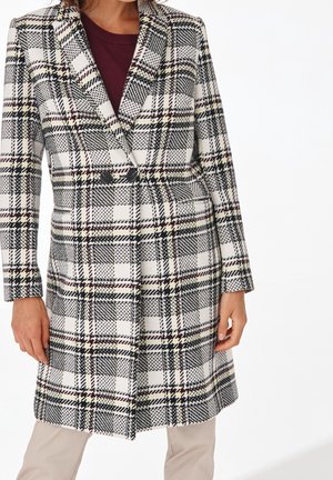 Kvinde iført knælængde hvid frakke med sort, rød og gul ternet mønster, over en bordeaux top og beige bukser.