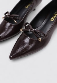 Zapatos de charol marrón con punta fina que presentan un tacón bajo, detalle de lazo en la parte frontal y acentos en tono dorado. Textura suave con un acabado brillante.