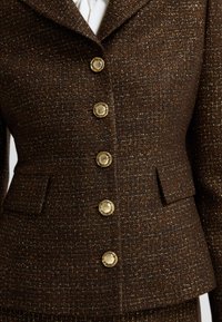 Brązowa marynarka w tweed z fakturowanym wykończeniem, z guzikami w kolorze złotym, z kołnierzem z wycięciem oraz dwiema przednimi kieszeniami.
