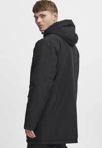 Chaqueta negra con capucha, hecha de un tejido suave, que presenta un corte relajado, mangas largas y un parche con el logotipo en el brazo izquierdo.