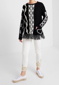 Black and white knitted sweater with fringe hem, patterned sleeves, and heart detail. Paired with white pants featuring gold embroidery and white sneakers.