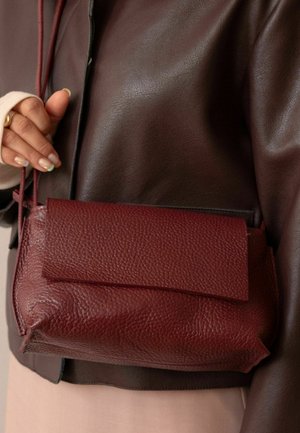 Mujer sosteniendo un bolso cruzado de cuero texturizado color burdeos, con chaqueta de cuero marrón oscuro y top beige, con la mano apoyada cerca de la correa del bolso.