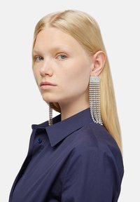 Orecchini argentati drammatici con strass a cascata e un perno rettangolare, indossati da un modello con una camicia colletto blu chiaro.