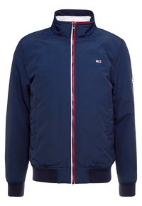 Marinblå bomberjacka med hög krage, vit foder, röda och vita dragkedjedetaljer, ribbade muddar och nederkant, med en liten logotyp på vänster bröst.