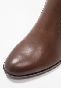 Hnědá kožená kotníková bota s hladkým povrchem, špičatou špičkou a jemnými švy. Disponuje nízkou černou podrážkou.