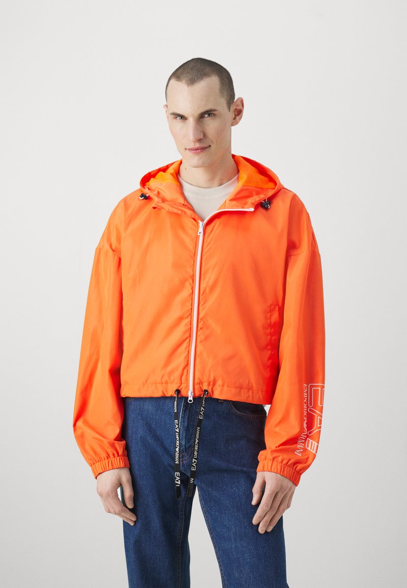 Lys orange vindjakke med hætte, elastiske manchetter, lynlåslukning, justerbart snor i kanten og logo på ærmet.