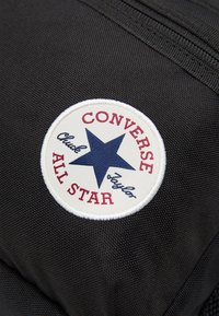 Zwarte stoffen tas met een ronde witte patch met een blauwe ster en rode opdruk "CONVERSE ALL STAR" en "Chuck Taylor." Gestructureerd oppervlak.