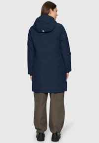 Woman wearing a navy blue hooded parka and brown wide-leg pants, standing facing away against a plain light gray background.