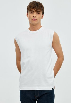 Joven de pie con las manos detrás de la espalda, vistiendo una camiseta blanca sin mangas y vaqueros oscuros contra un fondo claro y liso.