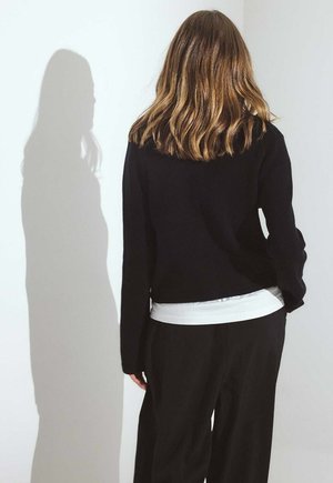 Suéter negro de canalé con mangas largas, superpuesto a una camisa blanca, combinado con pantalones negros. Textura suave visible; diseño sencillo.