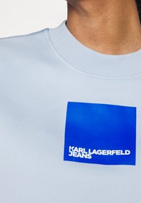 Jasnoniebieski bawełniany sweter z okrągłym dekoltem z niebieskim kwadratowym logo i białym napisem "KARL LAGERFELD JEANS" z przodu.