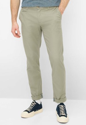 Man wearing rolled-up light beige pants, white socks, and black sneakers with white soles and laces, standing with one hand in pocket.