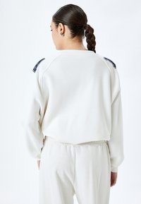 Maglione crema a maniche lunghe con maniche raglan. Presenta un collo a costine e dettagli in contrasto di colore blu navy sulle spalle. Texture morbida.