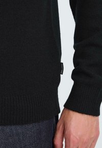 Zwarte gebreide trui met geribde mouwen en rand. Heeft een logotag aan de zijkant, gemaakt van zacht materiaal met een gladde textuur.