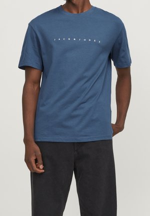 Mężczyzna w niebieskim t-shircie z krótkim rękawem z napisem „JACK & JONES” z przodu i czarnych spodniach, stojący na jednolitym, jasnym tle.