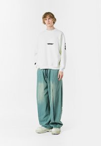Sudadera blanca de gran tamaño con texto negro y un bolsillo frontal, combinada con pantalones vaqueros de pierna ancha de color azul claro y zapatillas blancas gruesas.