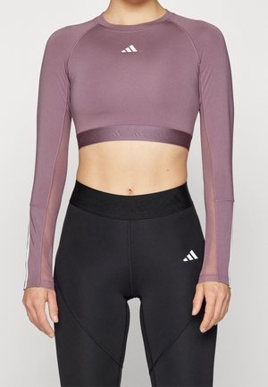 Kobieta ubrana w zamszowy, długi top sportowy z długim rękawem i czarne legginsy z wysokim stanem z logo Adidas na obu częściach odzieży.