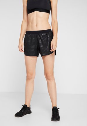 Kvinde iført sort sports-bh, mønstrede sorte atletikshorts og sorte sneakers stående med arme afslappet mod en ensfarvet baggrund.