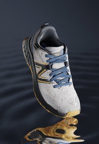 Šedá a bílá sportovní bota s texturovaným svrškem, modrými tkaničkami, černou podrážkou a zlatými akcenty, vystavená na reflexním povrchu.