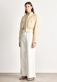 Sieviete valkājot bēša krāsas īsu žaketi, gaišu augstu apkakli, plašas baltas bikses un taupe krāsas apavus ar papēžiem, stāv uz neitrālas grīdas pret baltu sienu.