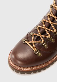 Brown leather hiking boot with yellow and brown patterned laces in metal hooks and rugged sole on white background.