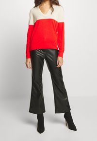 Svetr s červenobílým barevným blokováním a výstřihem do V, kombinovaný s černými flared kalhotami z umělé kůže a černými kotníkovými botami. Hladká textura.