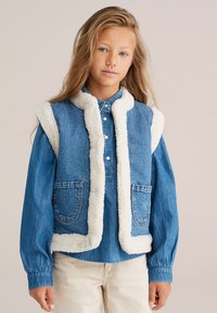 Gilet in denim con interno in pile bianco, colletto arrotondato e due tasche frontali. Indossato sopra una camicia blu con maniche a polsino.