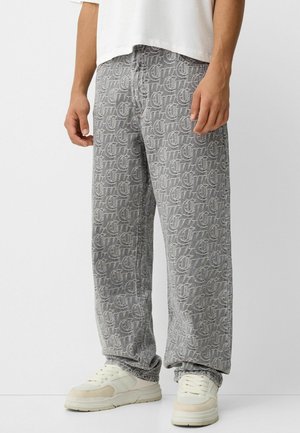 Osoba ubrana w szare szerokie spodnie z białym wzorem logo na całej powierzchni oraz kremowe sneakersy, stojąca na tle białego tła.