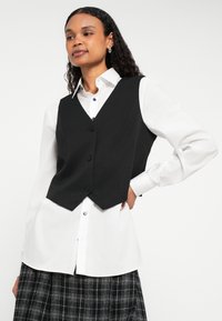 Čierna priliehavá vesta s výstrihom do V a tromi gombíkmi, nosená cez bielu košeľu so zapínaním na gombíky. Model má kučeravý účes.