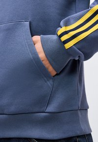 Χέρι τοποθετημένο στην τσέπη καγκουρό μπλε φούτερ με μανσέτα μανικιού με κίτρινη και μαύρη ρίγα, φορεμένο πάνω από σκούρο τζιν.