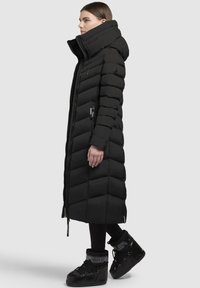 Juoda ilga pūkinė striukė su aukštu apykakle ir užtrauktuku. Suquiltuoti paneliai, šoninės kišenės ir derinama su juodais žiemos batais.