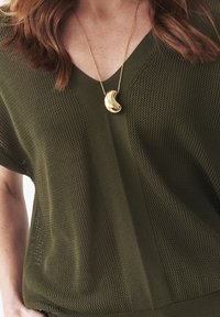 Olivově zelený pletený top s výstřihem do V, s texturovaným vzorem. Uprostřed visí zlatý náhrdelník v zakřiveném tvaru.