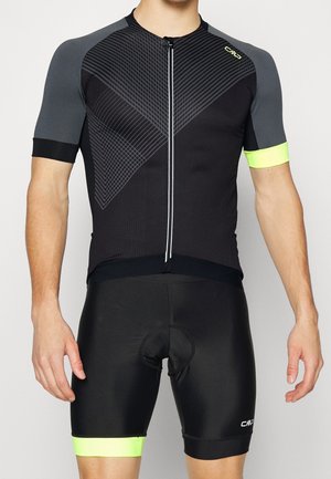 Miehen torso, jossa musta ja harmaa lyhythihainen pyöräilypaita neonkeltaisilla yksityiskohdilla ja siihen sopivat pehmustetut pyöräilyshortsit.
