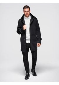 Homme portant un manteau noir sur un pull blanc, un pantalon noir et des bottes noires, se tenant devant un fond gris clair uni.