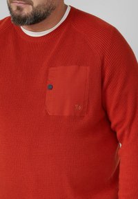Maglione rosso con texture e design a coste, dotato di una tasca sul petto e di un punto blu come accentuazione. Collo a giro con colletto interno bianco.