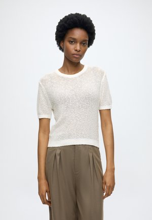 Young woman with short curly hair wearing a white textured short-sleeve top and loose brown high-waist trousers standing against a white background.