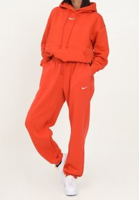Sudadera roja de Nike con un logo blanco, que cuenta con un bolsillo frontal, y pantalones de chándal rojos a juego con una cintura y puños elásticos. Zapatillas blancas.