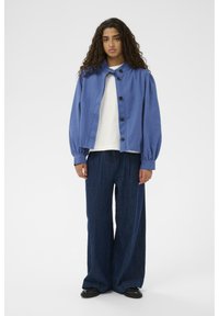 Modra bombažna jakna z balonastimi rokavi in črnimi gumbi, v kombinaciji s širokimi temnimi kavbojkami. Gladka tekstura, preprost dizajn.