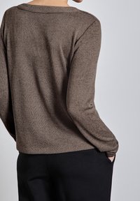 Ruskea, teksturoitu neulepaita, pyöreällä pääntiellä ja pitkillä hihoilla, yhdistettynä mustiin housuihin. Sileä kangas ja rento istuvuus ovat nähtävissä.