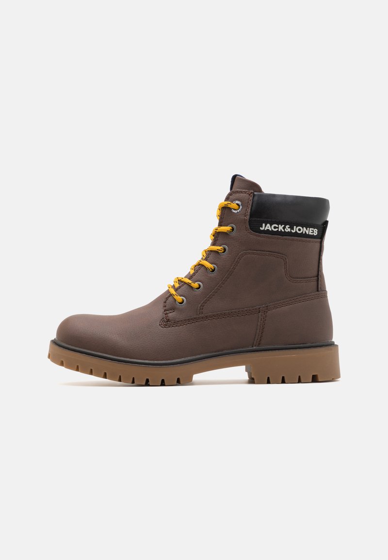 Bota de cuero marrón hasta el tobillo con cordones amarillos, cuello negro y suela de goma con relieve. Presenta un parche con el logo en el costado.