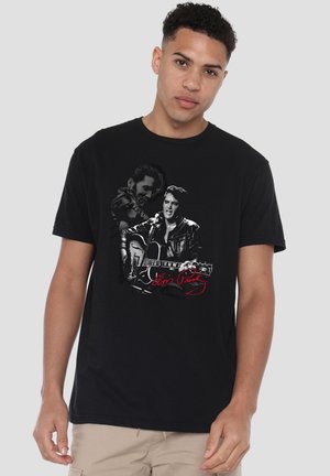Young man wearing black T-shirt featuring two black-and-white portraits of Elvis Presley, one playing guitar and singing, with red signature text.