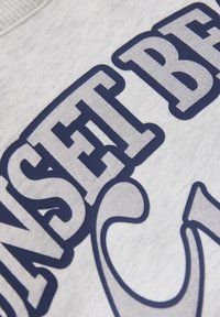 Grå sweatshirt med en mjuk textur, med fet marinblå text som säger "SUNSET BEACH" i en stor, stiliserad font.